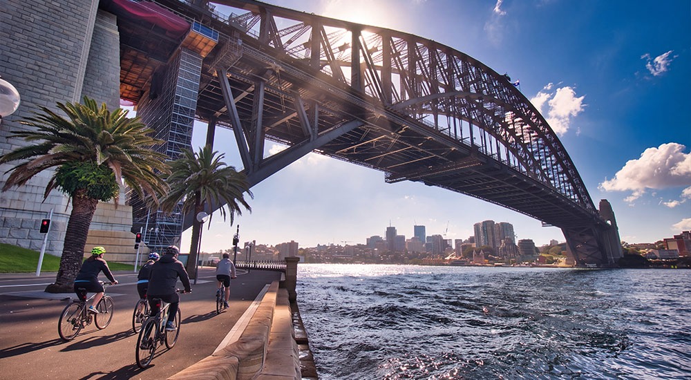 Cyclists on a guided Sydney bike tour riding along the waterfront beneath the Sydney Harbour Bridge on a sunny day