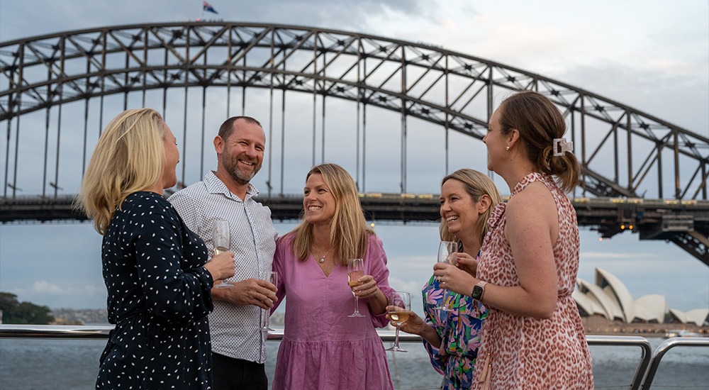 A group enjoying drinks and socialising on a Sydney Harbour lunch cruise, with the Sydney Harbour Bridge and Opera House in the background