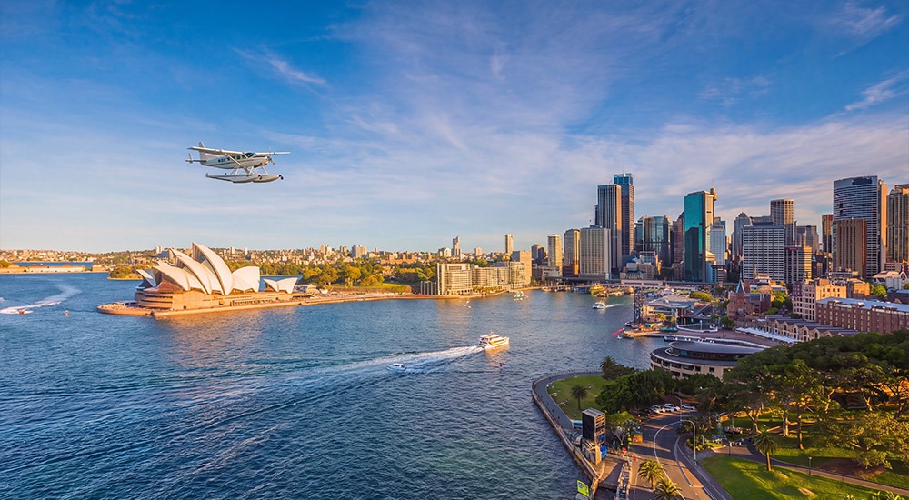 A seaplane flying over Sydney Harbour, with the Sydney Opera House, waterfront and city skyline visible in the afternoon sunlight.